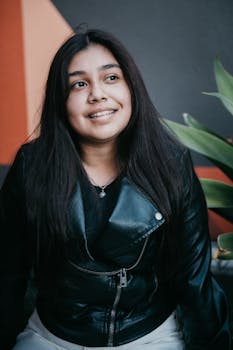 Portrait of a young woman in a leather jacket smiling outdoors with green foliage.