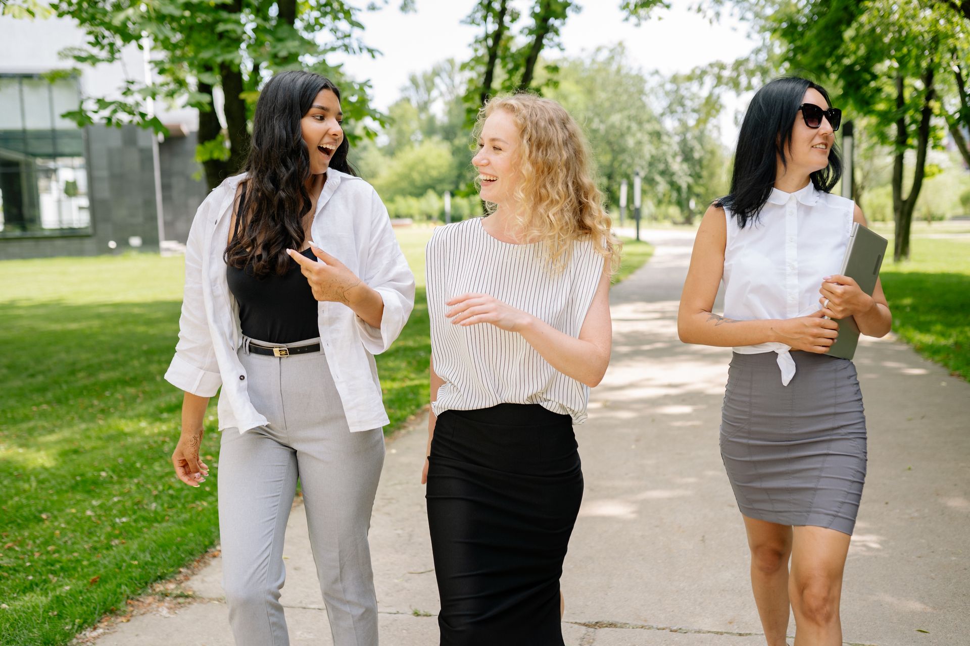 Three smiling businesswomen walk and converse in a sunny park setting, showcasing diversity and professional camaraderie.