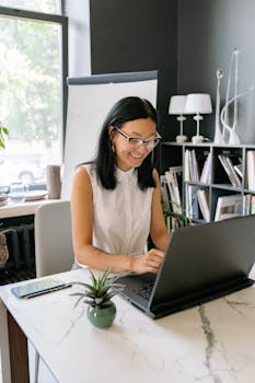 Smiling woman with glasses working on a laptop in a bright modern office setting.