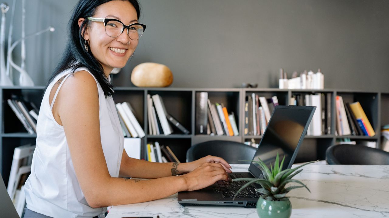 Cheerful businesswoman using laptop at a desk in a stylish office setup with bookshelves.