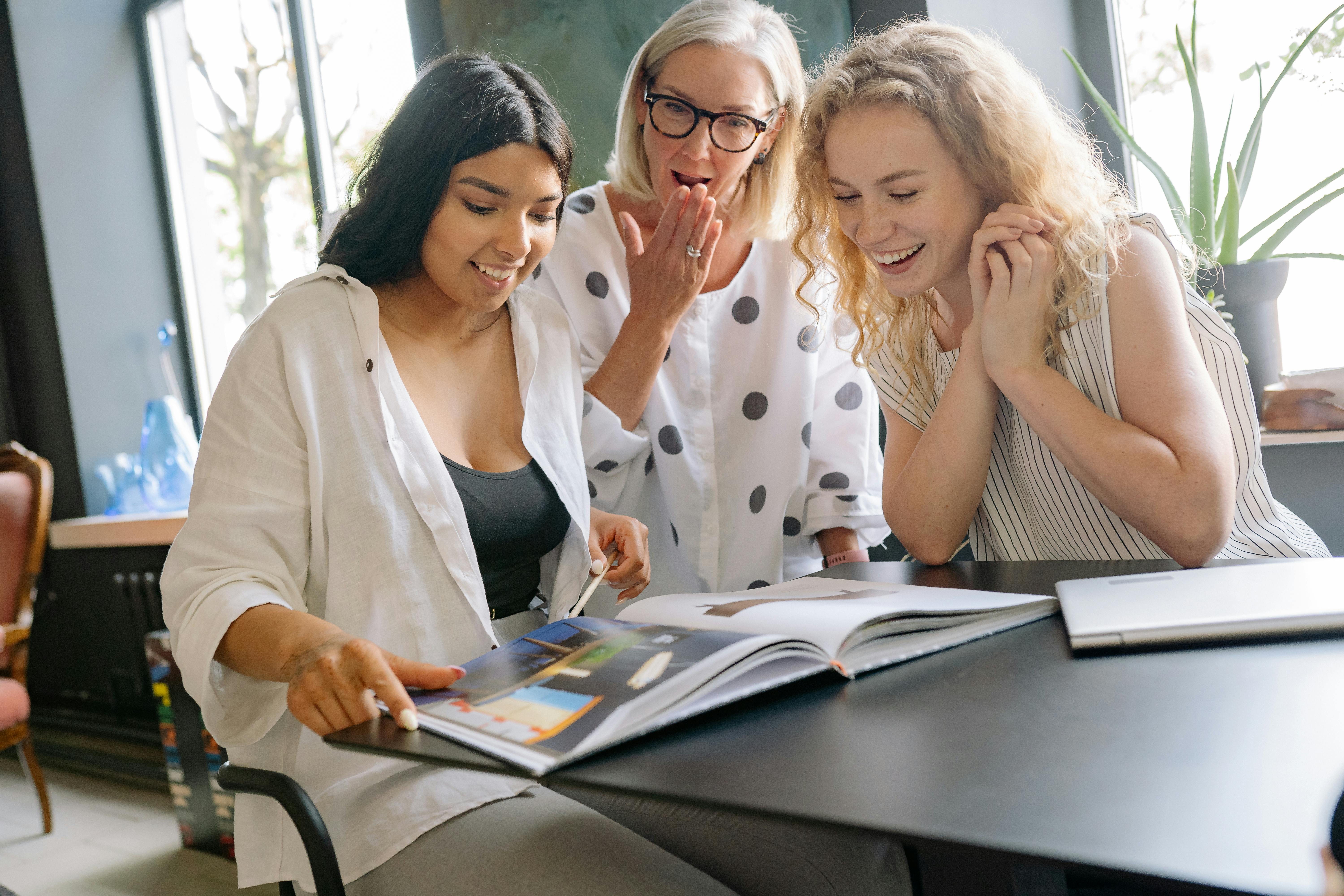 Three diverse women happily review a magazine in a bright office setting.