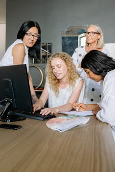 A diverse group of women discussing a project around a computer in a modern office setting.