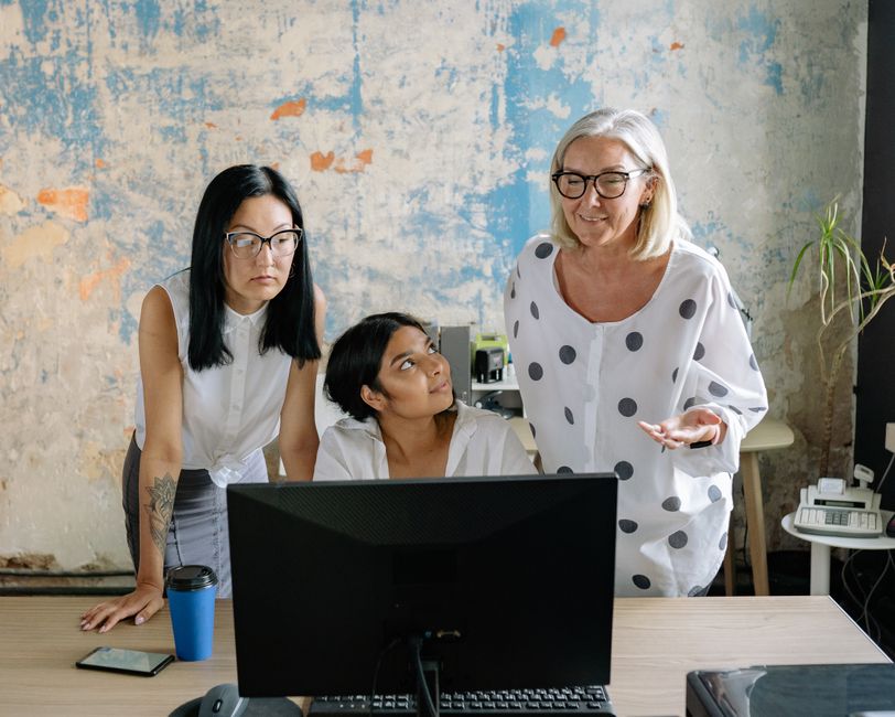 Três empresárias diversas colaborando em uma mesa em um ambiente de escritório moderno, focadas na tela do computador.