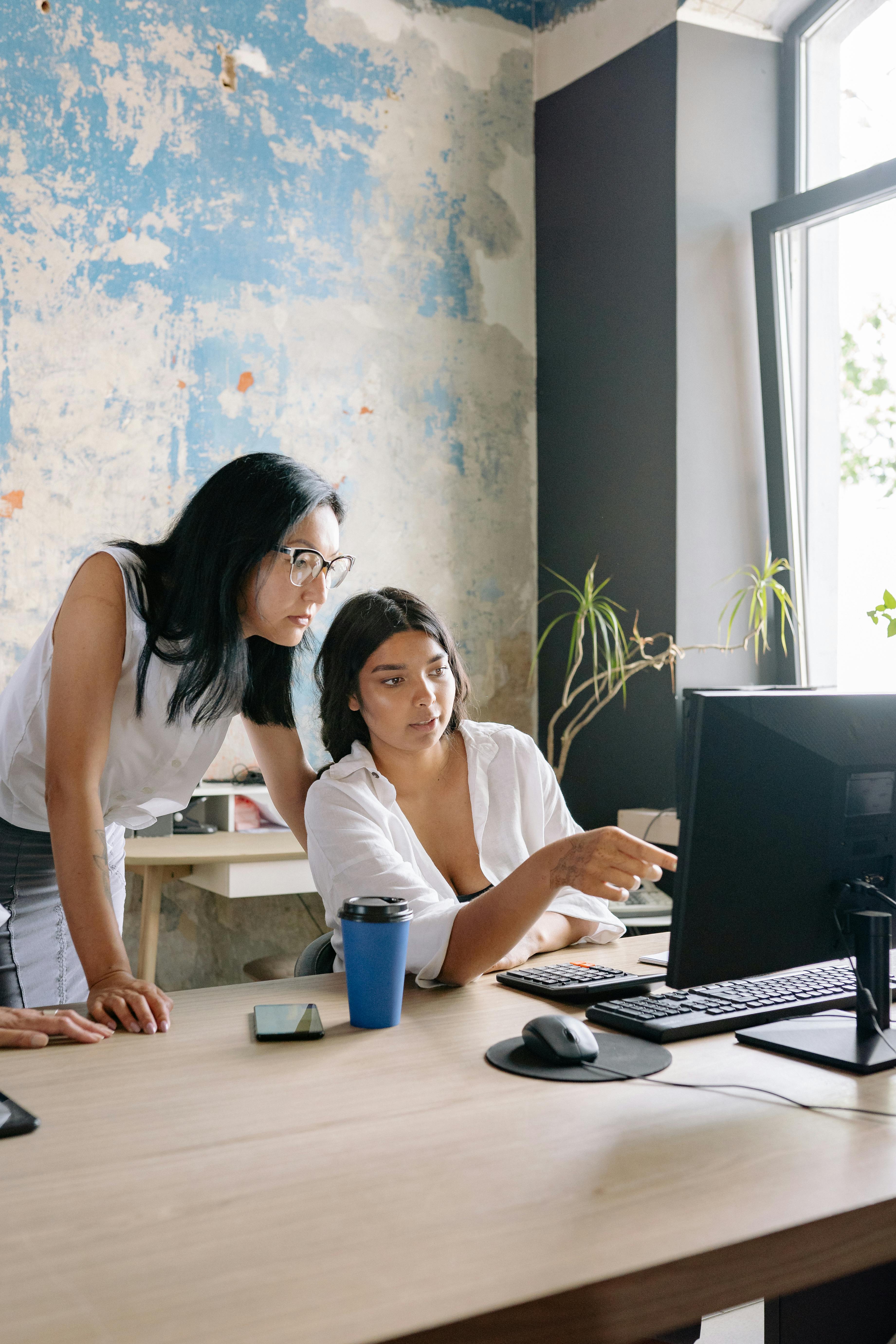 Two women working together in a modern office with a computer, showcasing teamwork and productivity.