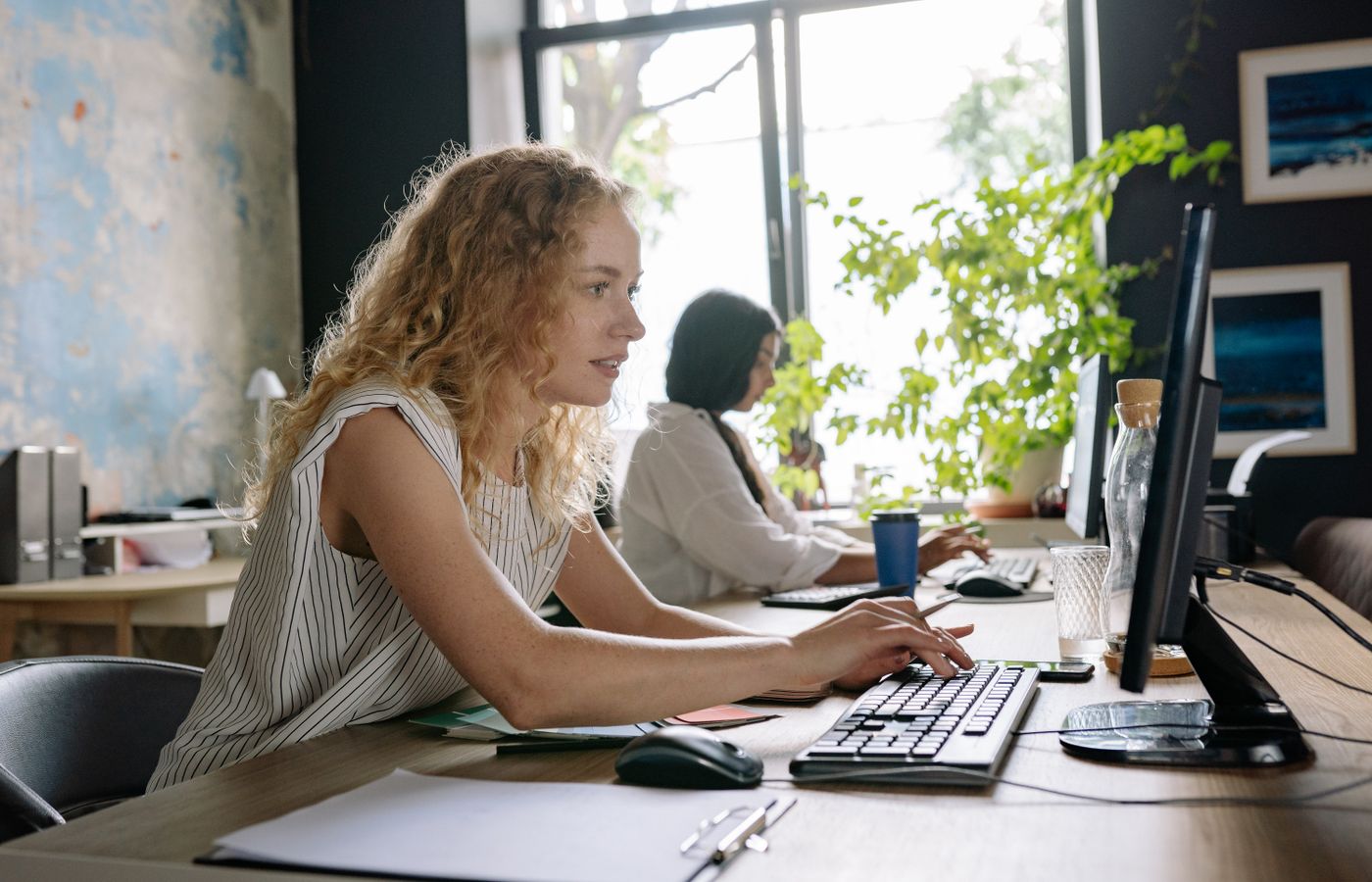 Two focused women working at a bright and modern office space, typing on computers at a shared desk.