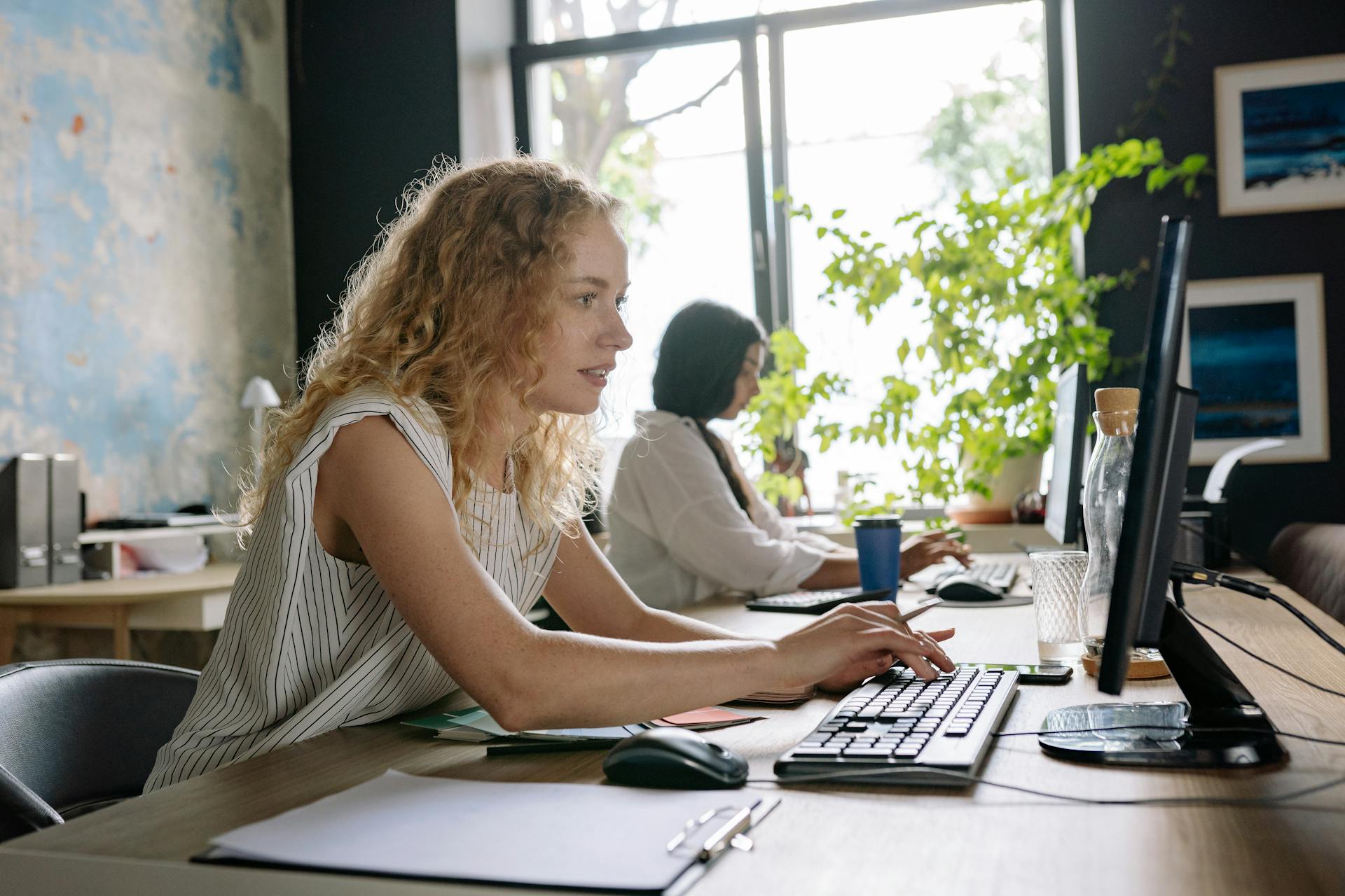 Two focused women working at a bright and modern office space, typing on computers at a shared desk.