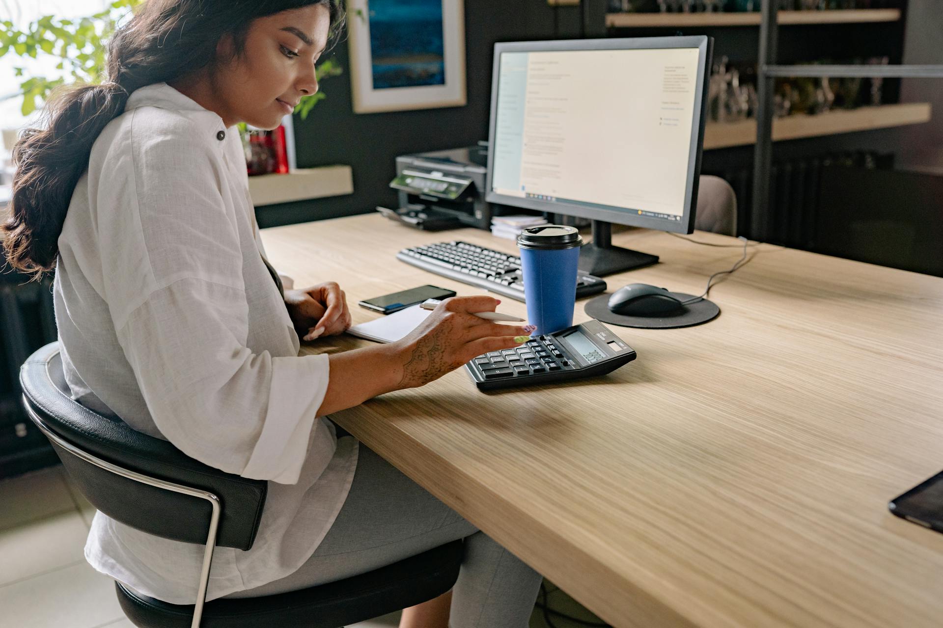 A professional woman using a calculator and computer at an office desk, focused on her work.