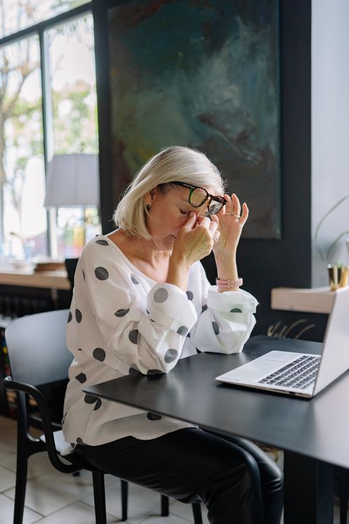Free A tired businesswoman in a polka dot blouse sits at a café by her laptop, rubbing her eyes. Stock Photo