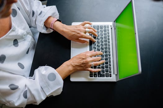 Overhead view of hands typing on laptop with green screen, polka dot shirt in focus.