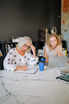 Two professional women working together in a modern office environment with laptops and documents.