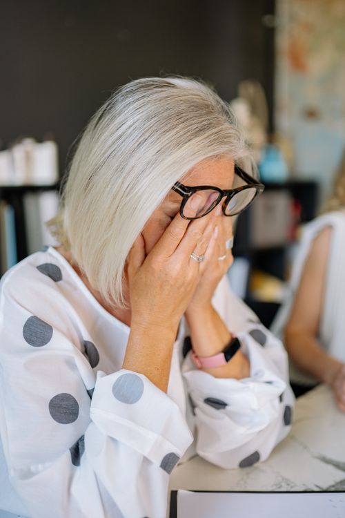 Free Middle-aged woman with eyeglasses in polka dot blouse showing stress indoors. Stock Photo