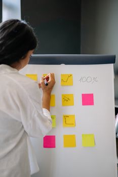 A woman writing on colorful sticky notes on a flip chart in an office environment.