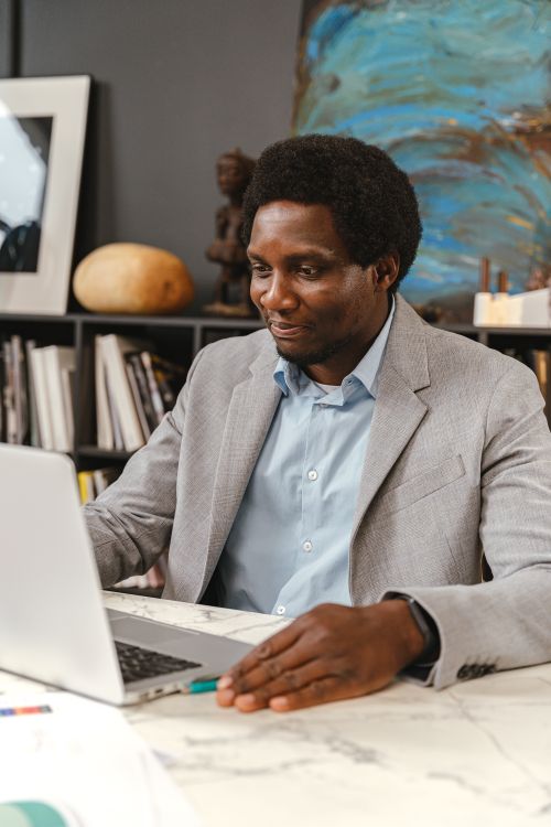 Free An adult man in a gray suit working on a laptop in an office environment, smiling and engaged. Stock Photo