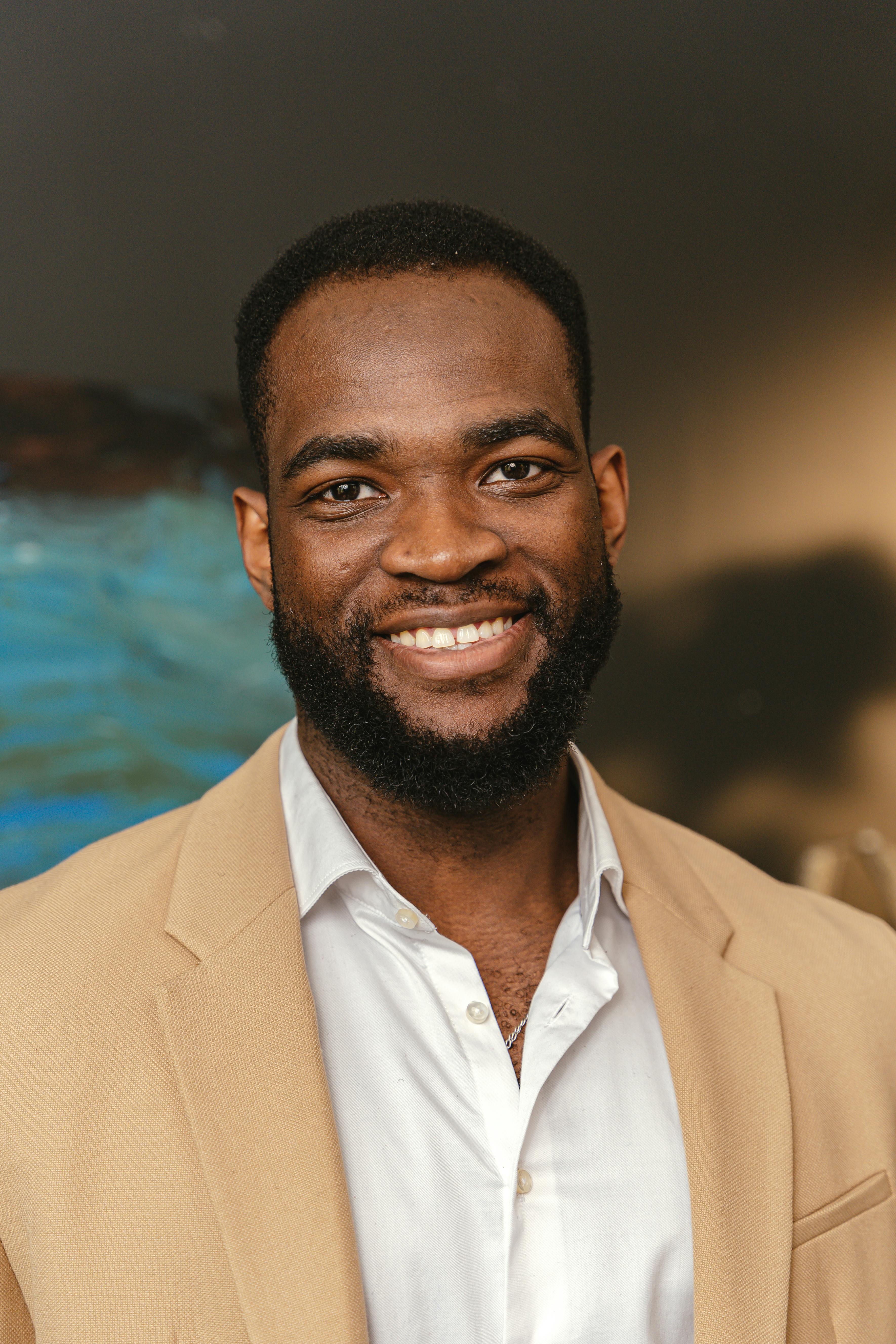 Portrait of a smiling black man with facial hair, indoors, wearing a beige blazer and white shirt.