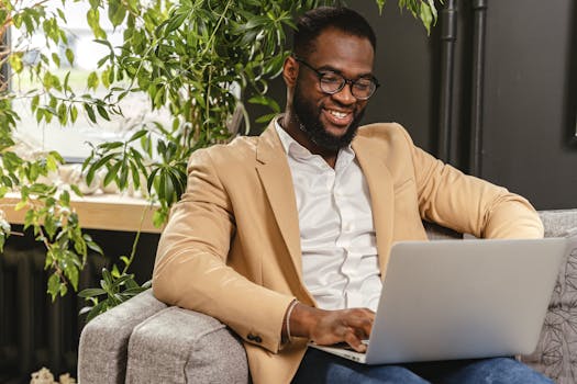 A man sitting on a couch works remotely on his laptop, smiling in a bright indoor setting.