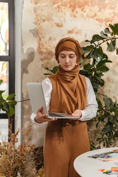 Woman in hijab holding a laptop in a rustic styled modern office with plants.