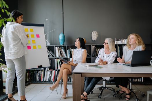A diverse group of women collaborating in an office setting, discussing ideas using a flip chart and laptop.