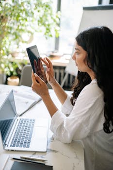 A focused woman in a bright office space using a tablet and laptop for business work.