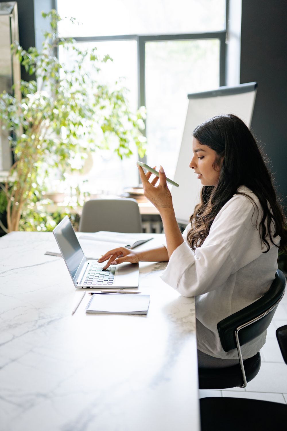 Woman using a laptop while holding a cellphone, managing communications while working remotely