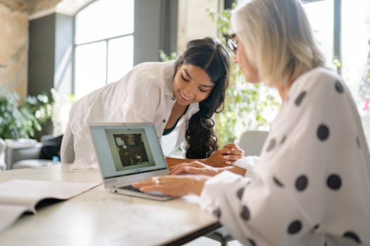 Two women collaborating in a bright, modern office with a laptop and green plants around.