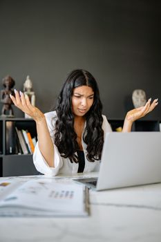 A frustrated businesswoman sits at her office desk, puzzled over her laptop.
