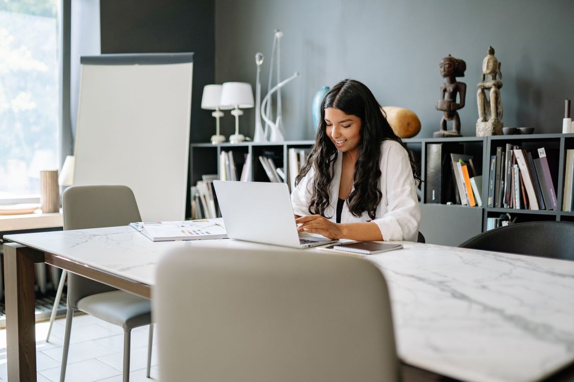 Smiling business owner typing prompts into a laptop at a marble table in a bright office