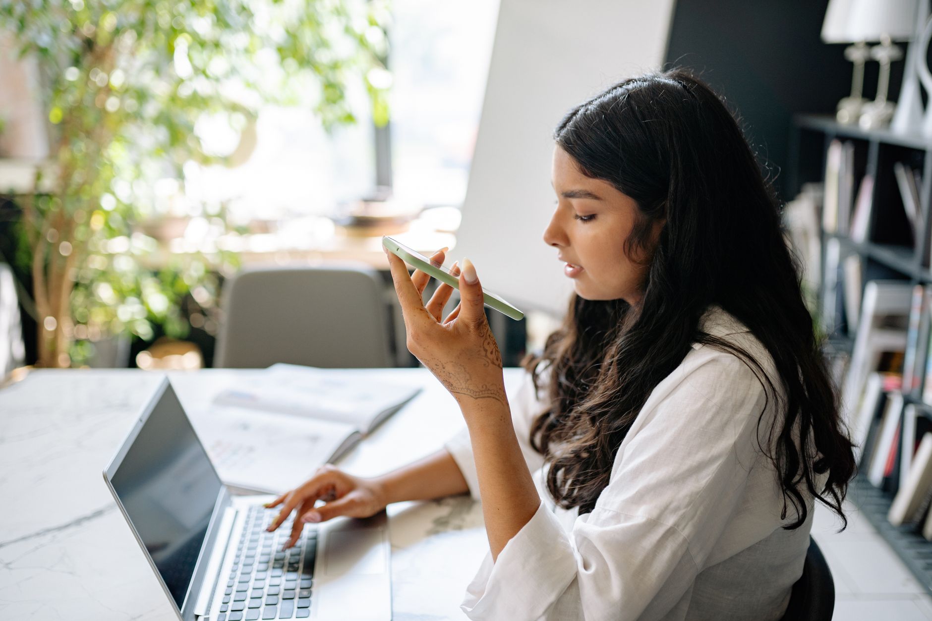 A focused woman uses her phone and laptop in a bright, home office setting.