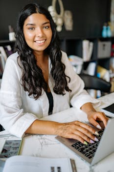 Confident young woman working on a laptop at home office desk, smiling at the camera.