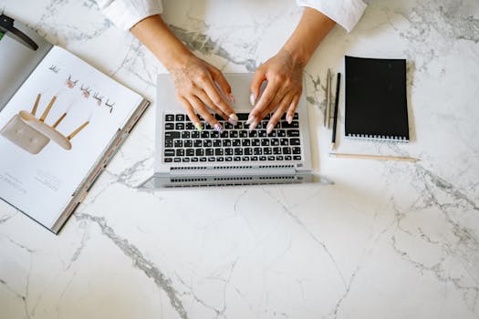 Close-up of hands working on a laptop, surrounded by a notebook, magazine, and pens on a marble desk.