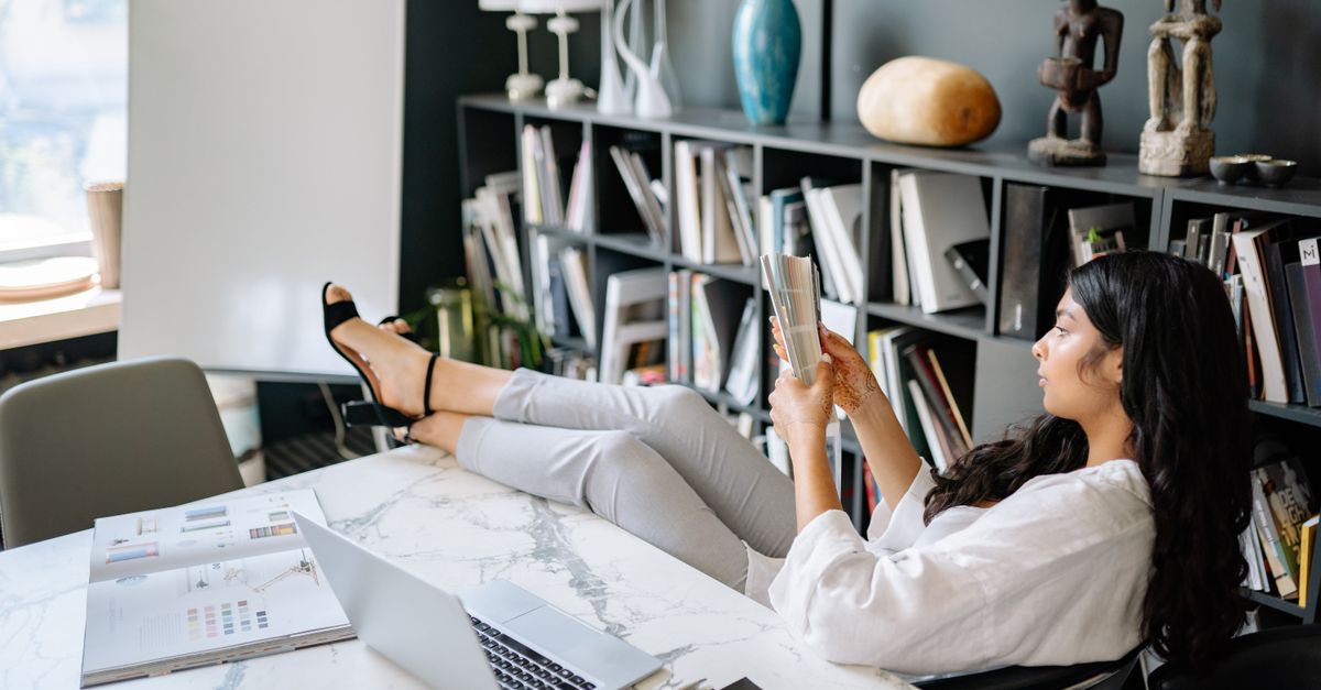 Woman reading a book in a modern office with laptop and papers, projecting a relaxing work environment.