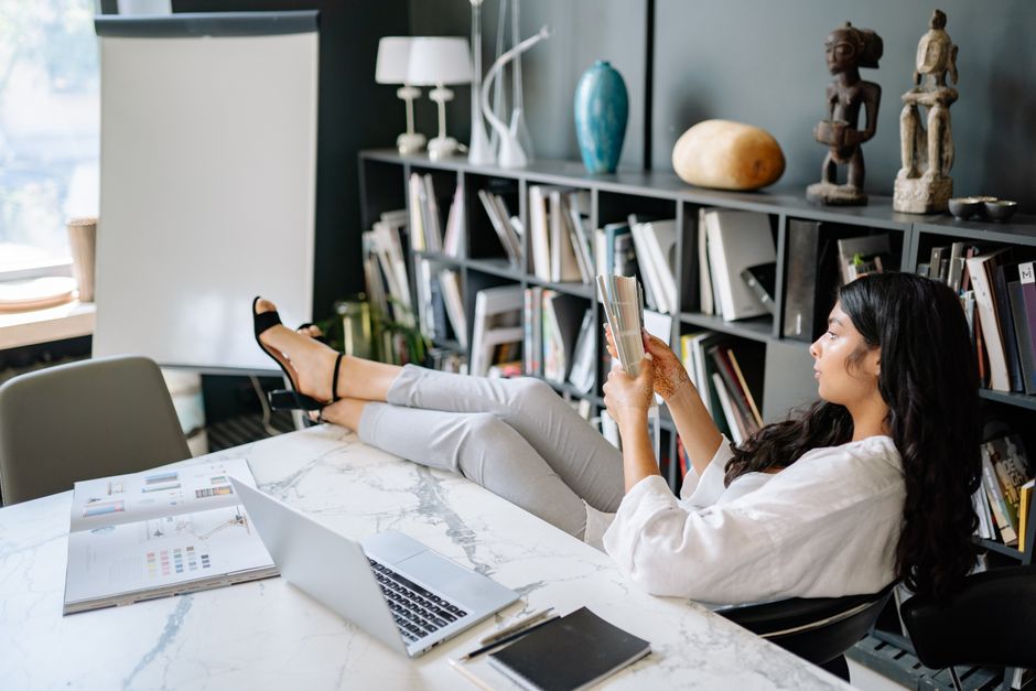 Woman reading a book in a modern office with laptop and papers, projecting a relaxing work environment.