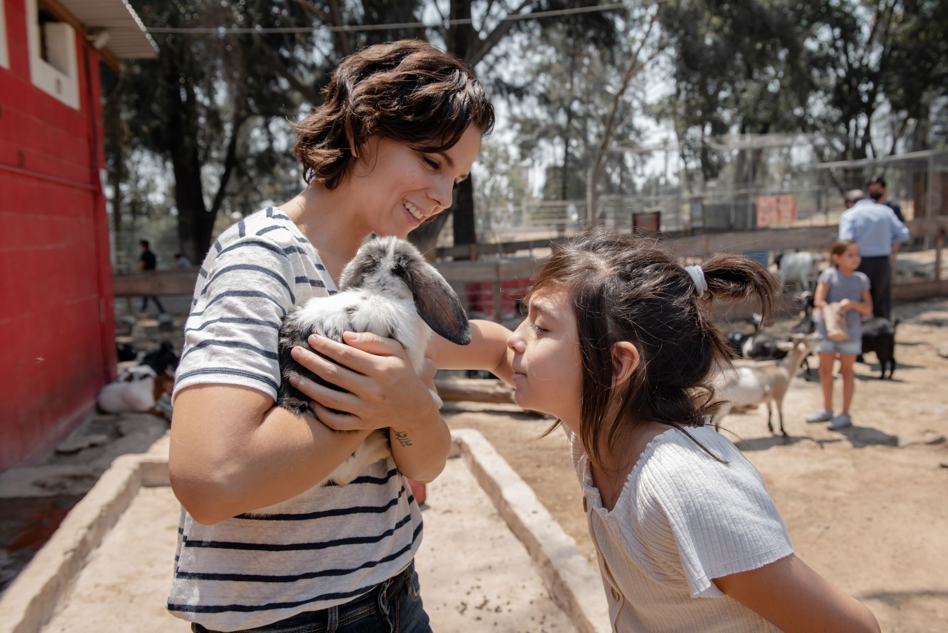 Mother and daughter bonding at an outdoor petting zoo, holding a rabbit on a sunny day.