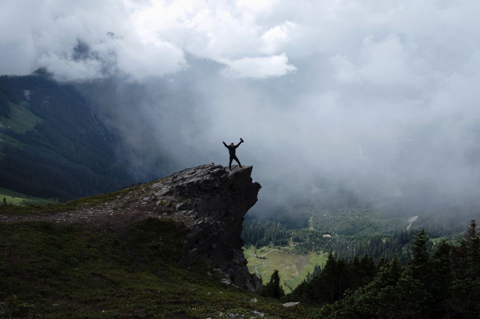 A lone hiker triumphantly stands on a misty mountain cliff, surrounded by clouds and forests.