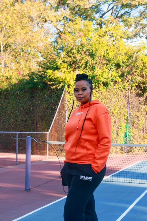 Free African American woman in vibrant orange hoodie posing at an outdoor tennis court. Stock Photo