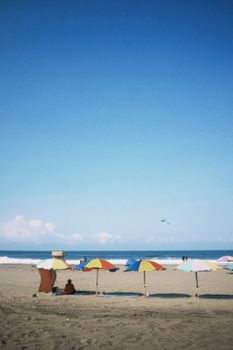 Vibrant beach umbrellas on a sandy shore under a clear blue sky with a distant kite flying.