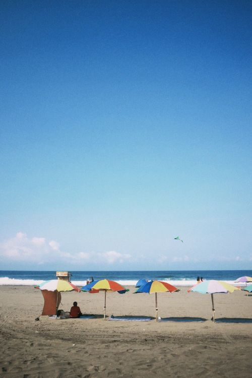 Free Vibrant beach umbrellas on a sandy shore under a clear blue sky with a distant kite flying. Stock Photo