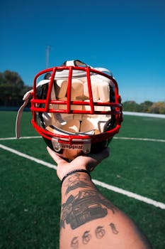 Close-up of a football helmet held by tattooed arm on a sunny sports field.