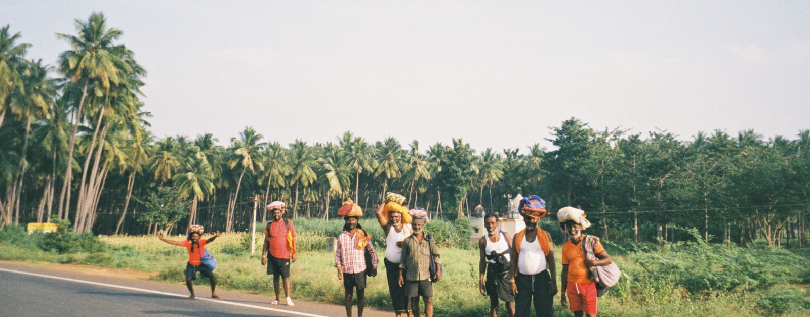 A diverse group of adults walking on a road surrounded by palm trees, carrying bundles on their heads.