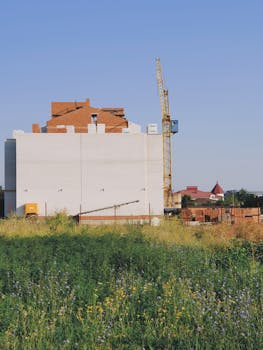 Construction site with crane and building in a lush green meadow under a clear blue sky.