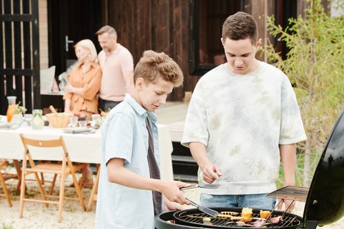 Free Two boys grilling at a family barbecue with parents in the background. Stock Photo