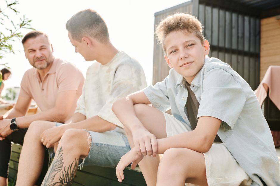 A cheerful boy sits outdoors with family enjoying a sunny day.