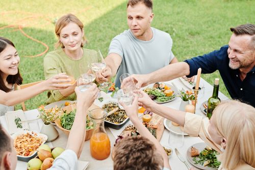 Free A joyful family with friends sharing a meal, toasting with drinks in a sunny outdoor setting. Stock Photo