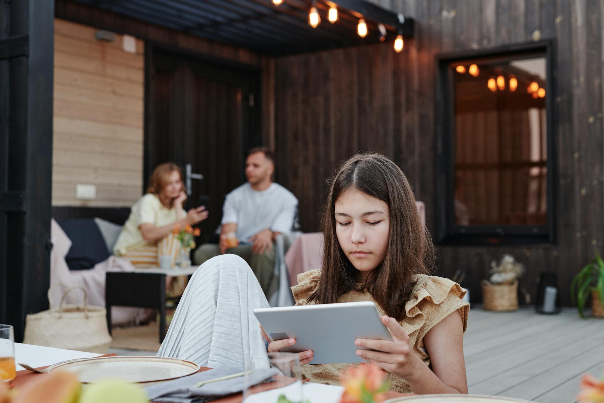 Teen girl focused on a tablet while sitting on a cozy outdoor patio with family in the background.