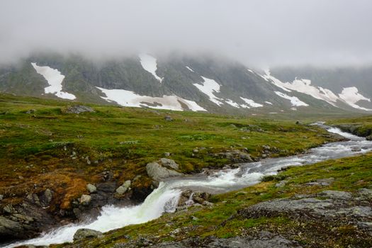 A tranquil foggy mountain valley with a flowing river and snow patches.