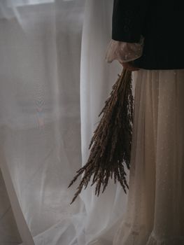Artistic close-up of a person holding a vintage dried grass bouquet next to sheer curtains.