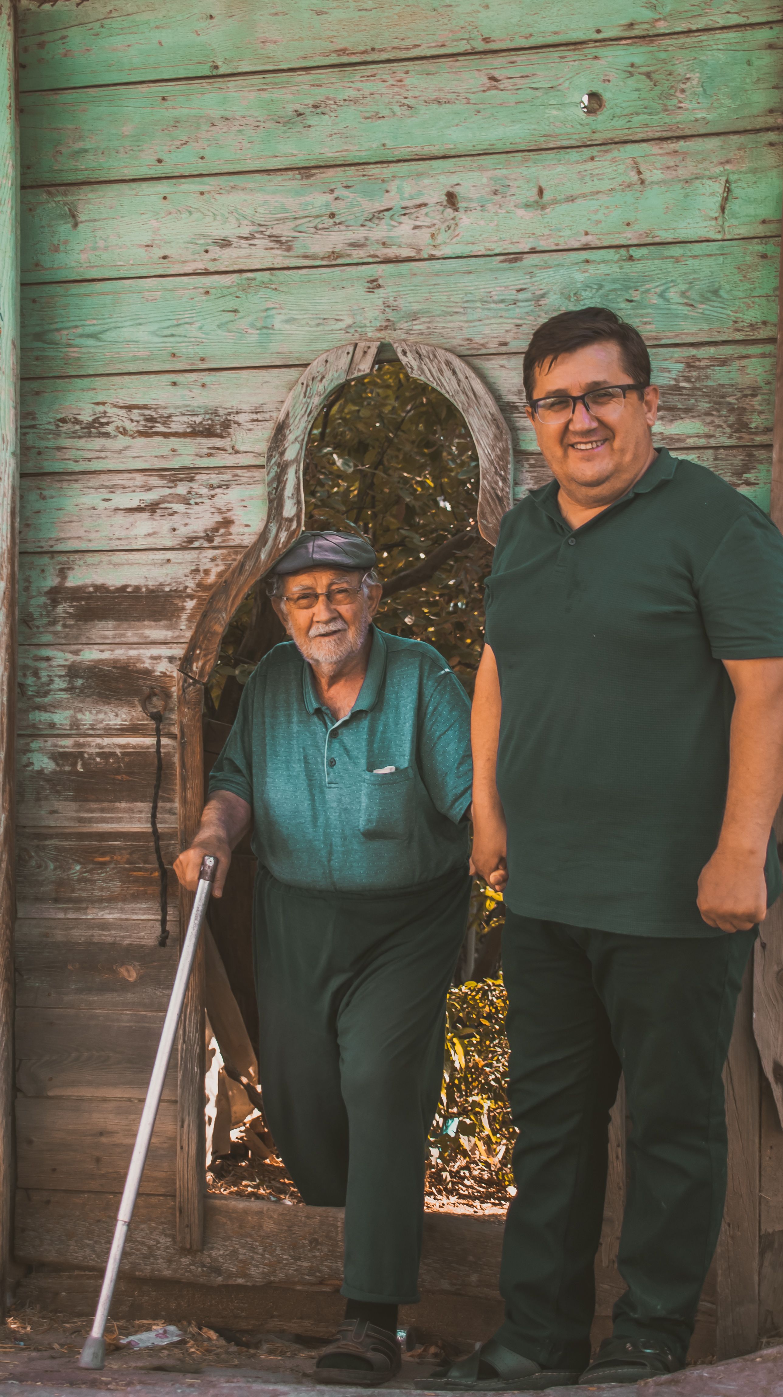 Elderly man with cane and supportive companion by a rustic wooden entrance, smiling warmly.