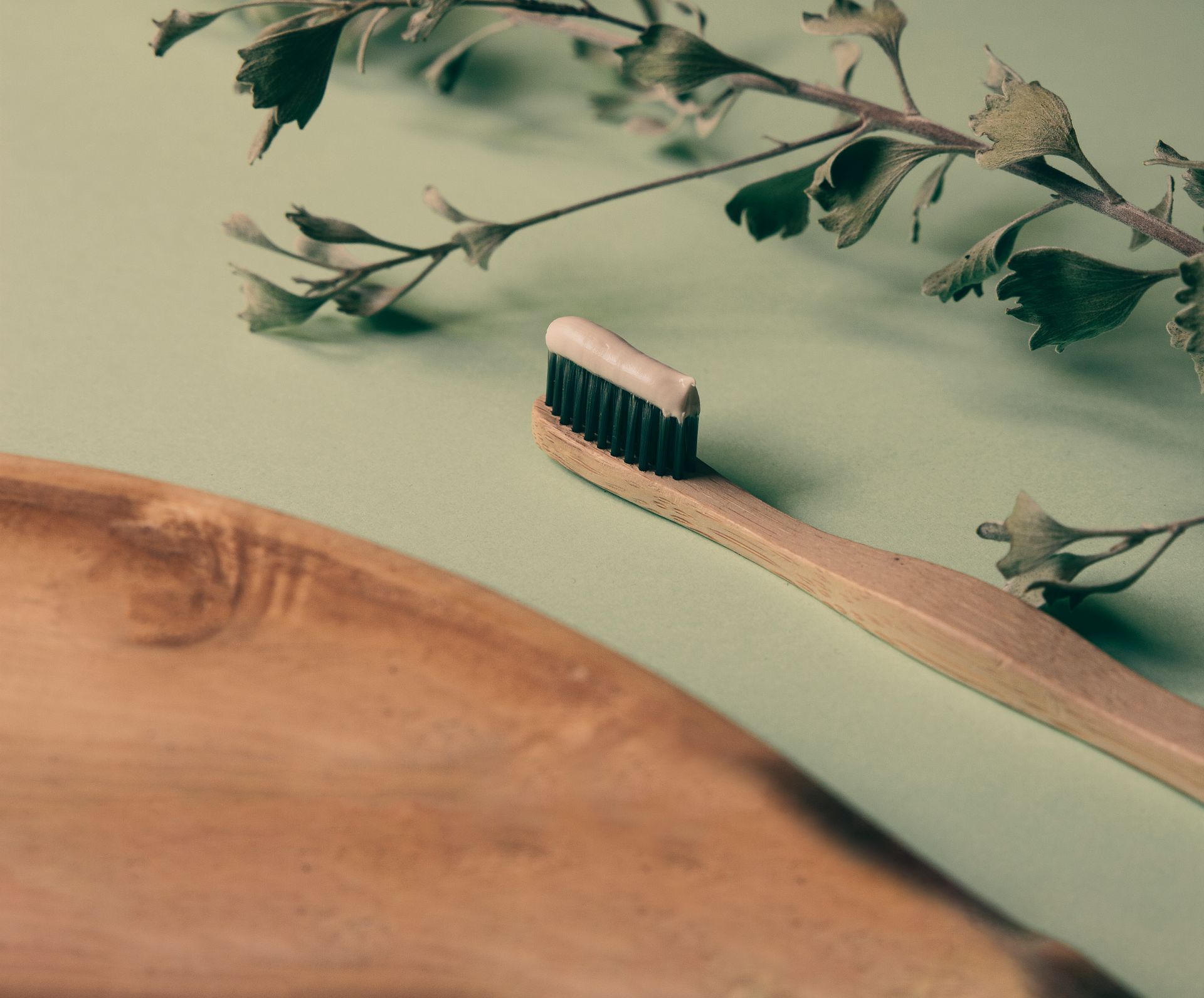 A bamboo toothbrush with toothpaste on a wooden tray beside green leaves, promoting natural oral care.