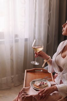 Woman enjoying pancakes and white wine on a wooden tray by the window.