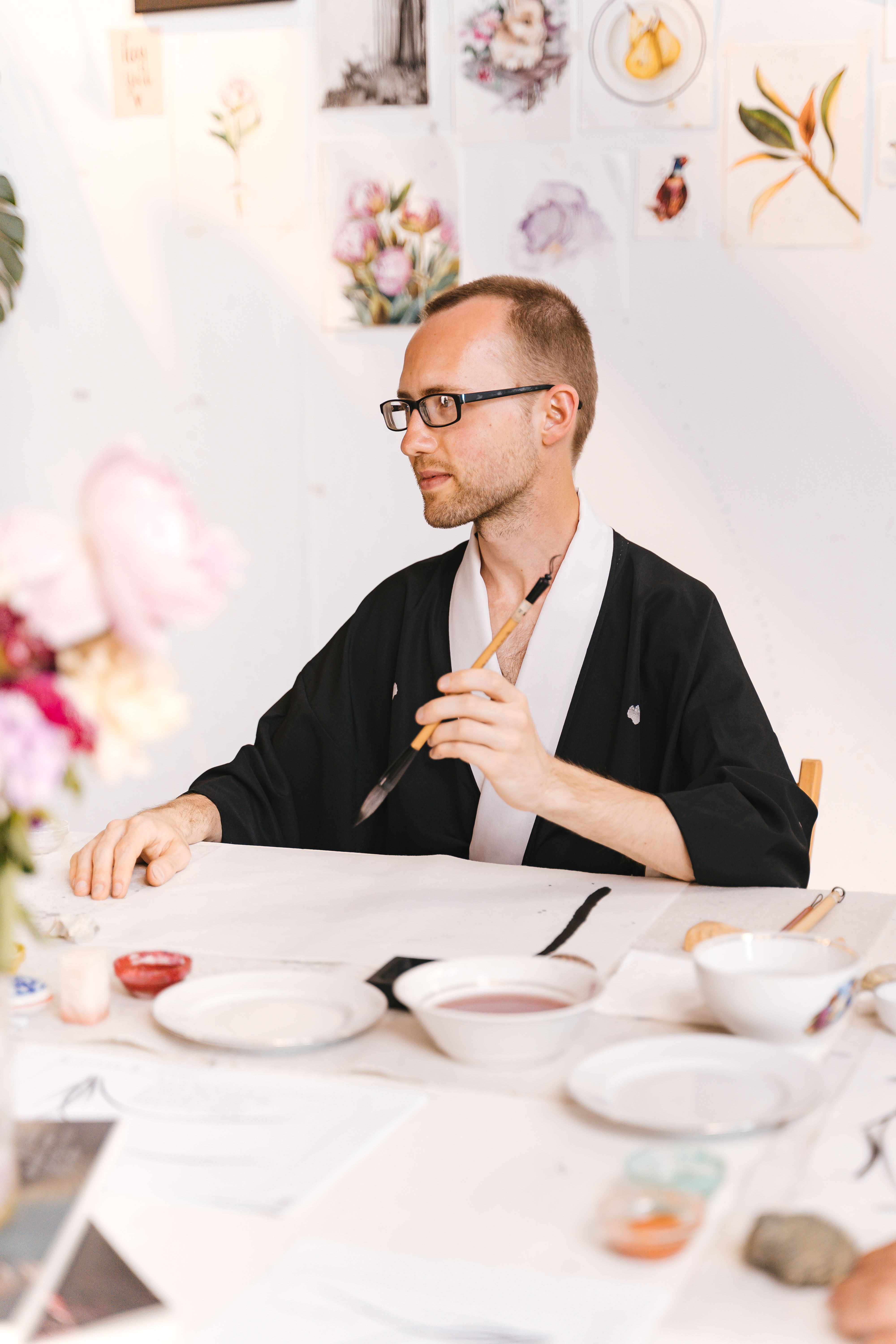 A man in a kimono focuses on calligraphy amidst an artistic, creative workspace.