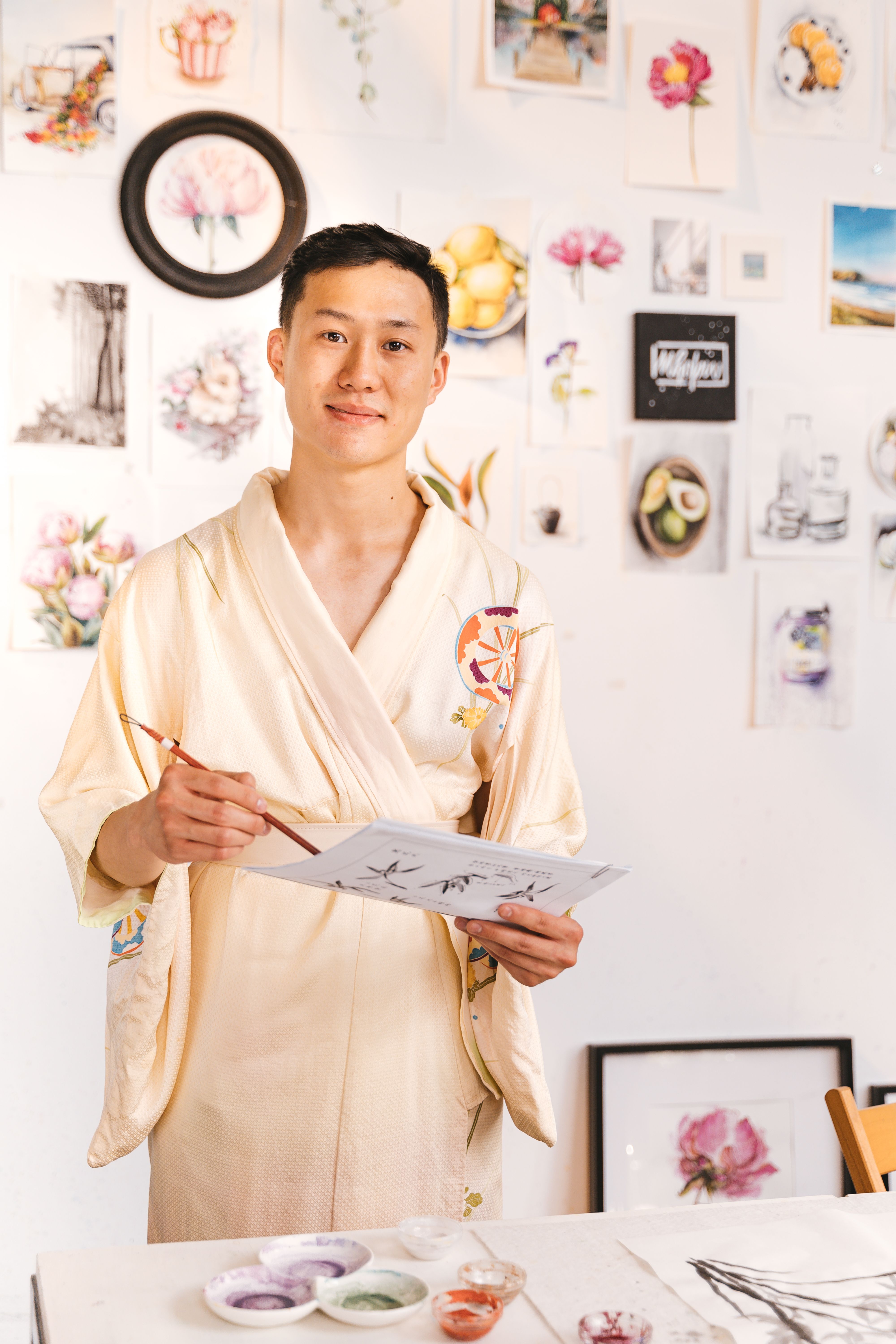 A creative Asian artist in a robe painting in a bright studio with various artworks.